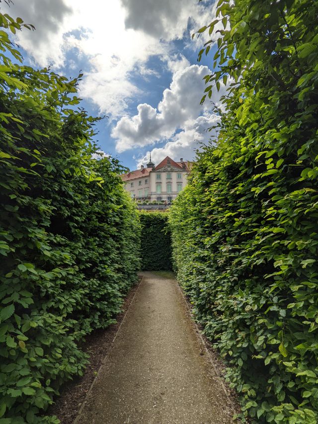 Schmaler Gartenweg zwischen hohen, dicht gewachsenen grünen Hecken mit historischem Gebäude im Hintergrund.